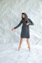 Load image into Gallery viewer, Wearing the Mitchley Long, a fairytale-like gray hooded coat, a dancer performs ballet on pointe against a white crumpled paper background. Her arms extend gracefully as she balances delicately on one foot, the woven cashmere flowing elegantly with her every move.