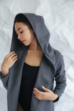 Load image into Gallery viewer, Wearing the Mitchley Long, a gray hooded coat with handmade snap buttons, a person in black stands against a crumpled white paper background, looking down while gently touching the edge of the hood.