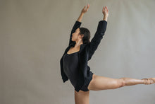 Load image into Gallery viewer, A dancer gracefully holds an arabesque pose against a neutral backdrop, wearing the Mornington unstructured jacket over a dark leotard. Her arms stretch overhead as one leg lifts elegantly behind her. The raw-edge stitches of the jacket add unique flair, enhancing the elegance of her movement.