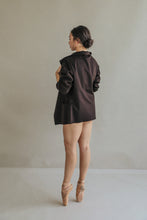 Load image into Gallery viewer, Wearing the Mornington, a long, unstructured jacket with raw-edge stitches, a person stands on pointe in ballet shoes, their hair styled in a bun as they face away against a plain and neutral-toned background.