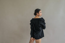 Load image into Gallery viewer, A person with dark hair stands against a neutral background, dressed in the oversized black Mornington shirt featuring raw-edge stitches. The person is slightly turned sideways, glancing over their shoulder.