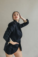 Load image into Gallery viewer, A person with long hair poses confidently in a minimalist setting, wearing the stylish "Sloane" dark blazer with structured shoulders. One hand is on their hip, while the other is raised, touching their head. The background is plain, highlighting their pose and fashion.