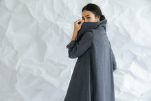 Load image into Gallery viewer, Wearing the Stamford Long cashmere coat, a person partially covers their face with its high collar while glancing over their shoulder. The backdrop features a crumpled white paper texture.