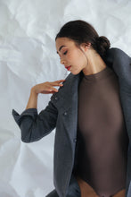 Load image into Gallery viewer, A woman poses gracefully against a crumpled fabric backdrop, wearing the Stamford Long coat. The dark cashmere coat, featuring an elegant collar, drapes over her fitted, long-sleeved top as she gazes downwards with her hair styled in a bun. Her right hand is elegantly lifted near her shoulder.