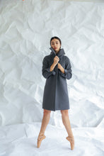 Load image into Gallery viewer, A woman in a Stamford Long B&W plaid coat stands on pointe in ballet shoes against a crumpled white paper backdrop, her hands adjusting the high collar while maintaining a poised and elegant posture.