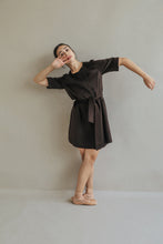 Load image into Gallery viewer, Someone wearing the Wexley, a dark brown, belted A-line dress, poses against a light gray wall. Standing on one leg with the other slightly bent, their arms suggest a dance pose, gracefully reminiscent of woven cashmere.