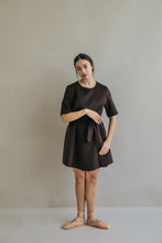 Load image into Gallery viewer, A woman models the Wexley, an A-line dark brown dress, against a plain backdrop. She pairs it with beige ballet shoes, has her hair tied back, and poses elegantly with one hand by her side and the other gently on her hip.