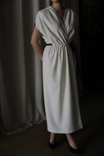 Load image into Gallery viewer, A person wearing the Lune sleeveless handmade white dress with a cinched waist stands with their hands placed behind their back. The figure is partially visible from the neck down to the feet, which are in dark-colored shoes, against a softly lit background.