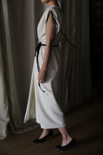 Load image into Gallery viewer, A person stands indoors wearing the Lune, a sleeveless, ankle-length handmade dress tied with a black belt. The back straps of the dress hang loosely as they are seen in profile, with black high heels on a wooden floor and cream-colored curtains in the background.