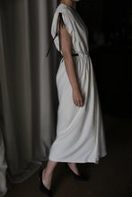 Load image into Gallery viewer, A person wearing the Lune, a flowing, sleeveless white raw silk dress with black ribbon detailing at the shoulders and around the waist, stands beside a curtain. Paired with black heels, this handmade dress encapsulates elegance in dim lighting, creating a soft and sophisticated atmosphere.