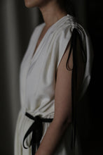 Load image into Gallery viewer, A person wearing the Lune dress, a white sleeveless raw silk creation featuring black ribbon details on the shoulders and waist, stands turned slightly to the side. The image captures only their upper body, emphasizing the intricate handmade details of the dress against a blurred background.