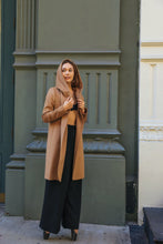 Load image into Gallery viewer, A woman in a fairytale-like Mitchley Long tan coat with a hood stands against an ornate building facade. Her woven cashmere ensemble pairs with black wide-leg trousers and heels, enhanced by handmade snap buttons, as she gazes thoughtfully to the side.