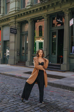 Load image into Gallery viewer, Wearing the Mitchley Long camel coat with handmade snap buttons over a black outfit, a woman confidently stands on a cobblestone street amid historic architecture, adding a fairytale charm to the urban scene.