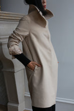 Load image into Gallery viewer, A person stands sideways wearing the Stamford Long cashmere coat, characterized by its high collar and hands in pockets. The coat is adorned with button details on the sleeves. The background is neutral, softly accented by a hint of a fireplace.