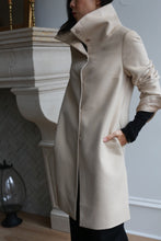 Load image into Gallery viewer, A person wearing a Stamford Long coat stands with hands in pockets next to a light-colored fireplace. The coat, crafted entirely from woven cashmere, features a dramatic high collar and is buttoned up. In the background, there is a wooden floor with a lantern near the fireplace.