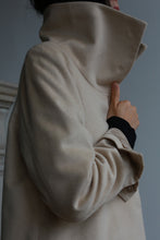 Load image into Gallery viewer, A person wearing the Stamford Long coat in light beige, featuring a dramatic high collar, is holding the collar with one hand. The texture of the coat, made from 100% woven cashmere, looks soft against a muted gray-blue background.
