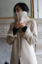 Load image into Gallery viewer, A person with dark hair is indoors, wearing a Stamford Long coat made of 100% woven cashmere. The dramatic collar is pulled up as they stand in front of a white stone wall, partially concealing their face.