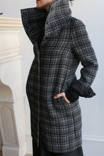 Load image into Gallery viewer, Indoors, a person wears the Stamford Long | Plaid coat in black and white cashmere, featuring a bold plaid pattern, large high collar, and pockets. Their face is partially turned away from the camera.