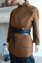 Load image into Gallery viewer, A person wearing a stylish brown coat crafted from genuine lambskin with an asymmetrical design and the Leather Obi with Grosgrain Ribbon around the waist stands indoors. The background features a decorative fireplace and vases. The person's face is not shown.