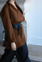 Load image into Gallery viewer, A person wearing a brown, genuine lambskin coat with wide lapels, cinched at the waist with a Leather Obi featuring a Grosgrain Ribbon tied in a bow, paired with black pants. The person is standing indoors near a white fireplace on light wooden flooring.