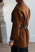 Load image into Gallery viewer, A person wearing a brown coat cinched with a Leather Obi with Grosgrain Ribbon stands facing away from the camera. The genuine lambskin coat accentuates their style, while dark hair tied back and a green bracelet add subtle details. The background features a white wall and a white stone fireplace, framing this chic scene perfectly.