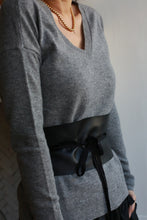 Load image into Gallery viewer, A person wears a gray V-neck sweater adorned with a gold chain necklace. A Leather Obi with Grosgrain Ribbon, made of genuine lambskin and featuring a tied bow, accentuates their waist. The background is a neutral-toned wall.