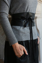 Load image into Gallery viewer, A person is wearing a gray, long-sleeved sweater with a Leather Obi featuring a Grosgrain Ribbon tied in a bow around the waist. They have one hand in the pocket of genuine lambskin pants or skirt. The background includes part of an interior with light-colored walls.
