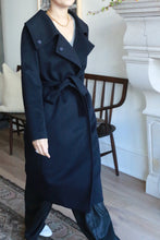 Load image into Gallery viewer, A person wearing the luxurious Malvern belted black coat stands indoors, partially obscured. They are in front of a wooden chair and a fireplace, with a patterned rug on the floor. A necklace and casual black pants complement the outfit, highlighting the woven cashmere's elegance.