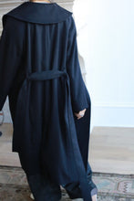 Load image into Gallery viewer, A person walks on a hardwood floor wearing the Malvern, a long, double-breasted luxury coat featuring a wide collar and belt. They are in a room with a light-colored wall, where part of a patterned rug is visible. The image captures the scene from behind.