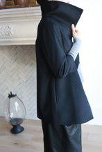 Load image into Gallery viewer, A person stands indoors wearing a Stamford, a stylish black cashmere coat with a high collar and 3/4 sleeves, partially turned up. The person's face is not visible. The background features a white, herringbone-patterned wall with decorative molding and a large decorative glass object on the floor.
