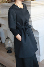 Load image into Gallery viewer, A person dons the stylish Wexley black wrap dress with an A-line cut, paired with black pants and a gold necklace. They stand indoors near a decorative fireplace and lantern, complemented by an elegant double-breasted coat. Their left hand rests in a pocket.