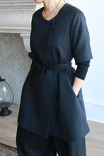 Load image into Gallery viewer, A person indoors, styled in the Wexley, a black woven cashmere coat with a three-quarter sleeve A-line cut and tied belt, paired with black pants. They accessorize with a gold necklace as they stand near a fireplace featuring herringbone design tiles in the background.