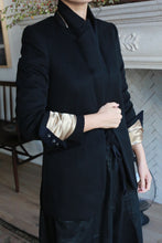 Load image into Gallery viewer, A person is seen from the shoulders down wearing the Sloane blazer in black, featuring structured shoulders and sleeves partly rolled up to reveal a satin gold lining. Their hand grasps the lapel of the blazer. A fireplace and part of a wooden chair are visible in the background.