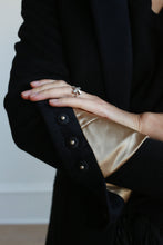 Load image into Gallery viewer, A person in a black Sloane equestrian-length coat with golden silk lining holds their hand against their chest, revealing a silver ring with a large, clear gemstone on their finger. The woven cashmere Sloane coat features three black buttons on the cuff and structured shoulders.