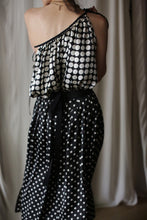 Load image into Gallery viewer, A person is wearing a stylish black and white polka dot one-shoulder dress with the Charmeuse Pleated Wrap Skirt | B/W Polkadot. The background features soft folds of light-colored fabric, creating an elegant and minimalist setting. The person's shoulder is adorned with layered gold necklaces.