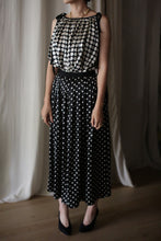 Load image into Gallery viewer, A woman is standing indoors wearing a black sleeveless dress with white polka dots and a fitted waistband. The Charmeuse Pleated Wrap Skirt | B/W Polkadot adds elegance, complemented by silk charmeuse fabric. She is accessorized with a necklace and black high-heeled shoes. The background features a neutral curtain and wooden flooring.