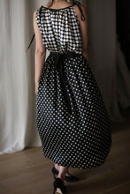Load image into Gallery viewer, A person wearing a Charmeuse Pleated Wrap Skirt | B/W Polkadot, tied with a black ribbon at the waist, stands indoors on a wooden floor. The image is captured from behind, revealing the graceful flow of the silk charmeuse fabric. The lower section of a curtain is visible in the background.