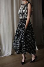 Load image into Gallery viewer, A person is dressed in a Charmeuse Pleated Wrap Skirt | B/W Polkadot, reminiscent of silk charmeuse. They are wearing black high heels and a gold chain necklace. The background features a white curtain, and the person is standing on a wooden floor. Their face is not visible.
