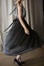 Load image into Gallery viewer, A woman in a sleeveless black dress with white polka dots twirls, making the skirt of her Charmeuse Pleated Wrap Skirt | B/W Polkadot flare out. She is wearing black high heels and stands on a wooden floor with white curtains in the background.