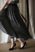 Load image into Gallery viewer, A person wearing a Charmeuse Pleated Wrap Skirt | B/W Polkadot twirls around, making the skirt flare out. The outfit, adorned with black high heels, flows elegantly against the backdrop of draped white curtains.