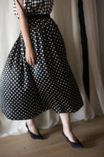 Load image into Gallery viewer, A person wearing a Charmeuse Pleated Wrap Skirt | B/W Polkadot and a matching sleeveless top walks across a room with light-colored curtains. They are also wearing black shoes. The image captures the movement of the flowing skirt and part of the person's arm and legs.