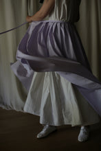 Load image into Gallery viewer, A person swirls in a dress with layers of white and lilac Typewriter Wrap Skirt | Amethyst. The movement creates dynamic folds in the wrap skirt, emphasizing the fluidity of the garment. The scene captures a sense of motion and elegance, perfect for summer plans. Only part of the person is visible, mainly the dress and legs.