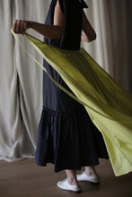 Load image into Gallery viewer, A person wearing a dark, sleeveless dress and white shoes is seen holding the ends of a large, flowing Typewriter Wrap Skirt | Citrine, creating a striking contrast against their outfit. The background features light-colored curtains.