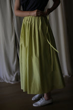 Load image into Gallery viewer, A person is shown from the waist down, wearing a long, pleated yellow Typewriter Wrap Skirt | Citrine and white shoes. They are adjusting the waistband of the skirt. The background features draped fabric in neutral tones.