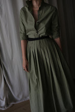 Load image into Gallery viewer, A person wearing the olive Typewriter Wrap Skirt with a black belt and pearl necklace stands before cream-colored curtains. The collared, buttoned long dress features a pleated skirt. Their face is not visible.