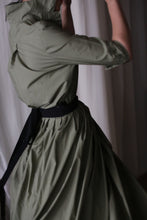 Load image into Gallery viewer, A person wearing an olive green Typewriter Wrap Skirt is captured mid-movement, partially turned away from the camera in soft lighting against a neutral background.