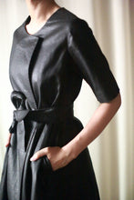 Load image into Gallery viewer, A person is wearing the Waxed Linen Wexley, a black, short-sleeve, belted dress with a textured, leather-like finish and two side pockets. The waxed linen design adds to its allure. With hands in the pockets and their head out of frame, the background remains neutral and out of focus. A truly versatile piece.