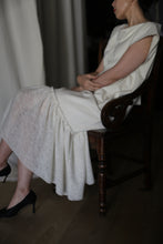 Load image into Gallery viewer, A person is sitting on the Paix wooden chair with their hands resting on their lap. They are wearing a textured, white dress featuring delicate lambswool ruffles and black high heels. The background appears to be neutral and subdued, consisting of fabric drapes. Their face is not fully visible.