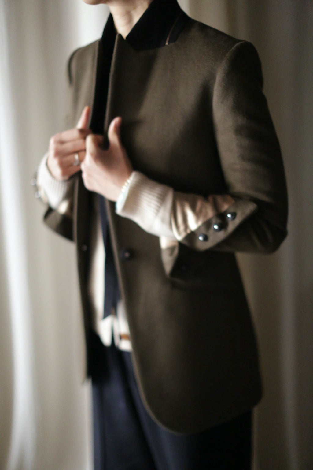 The Sloane | Olive / Black is an equestrian length olive green jacket with a black collar and black buttons on the cuffs, worn over a light-colored woven cashmere sweater.
