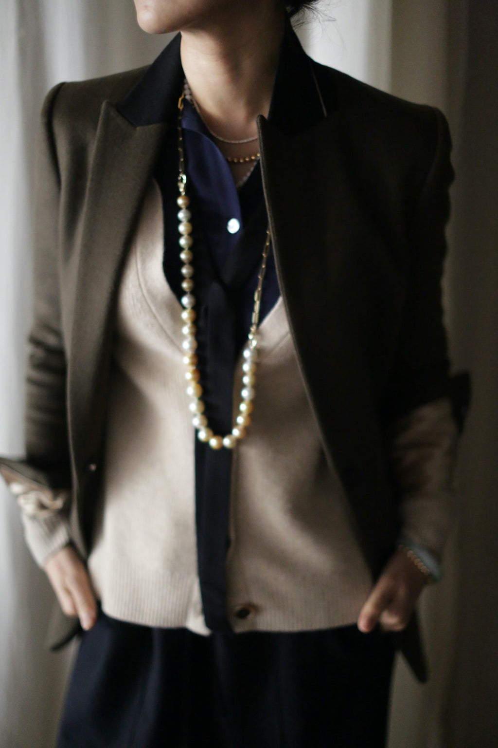 A person wears a Sloane | Taupe / Black tuxedo jacket over a woven cashmere cardigan and dark button-down shirt, accented by a long pearl necklace.